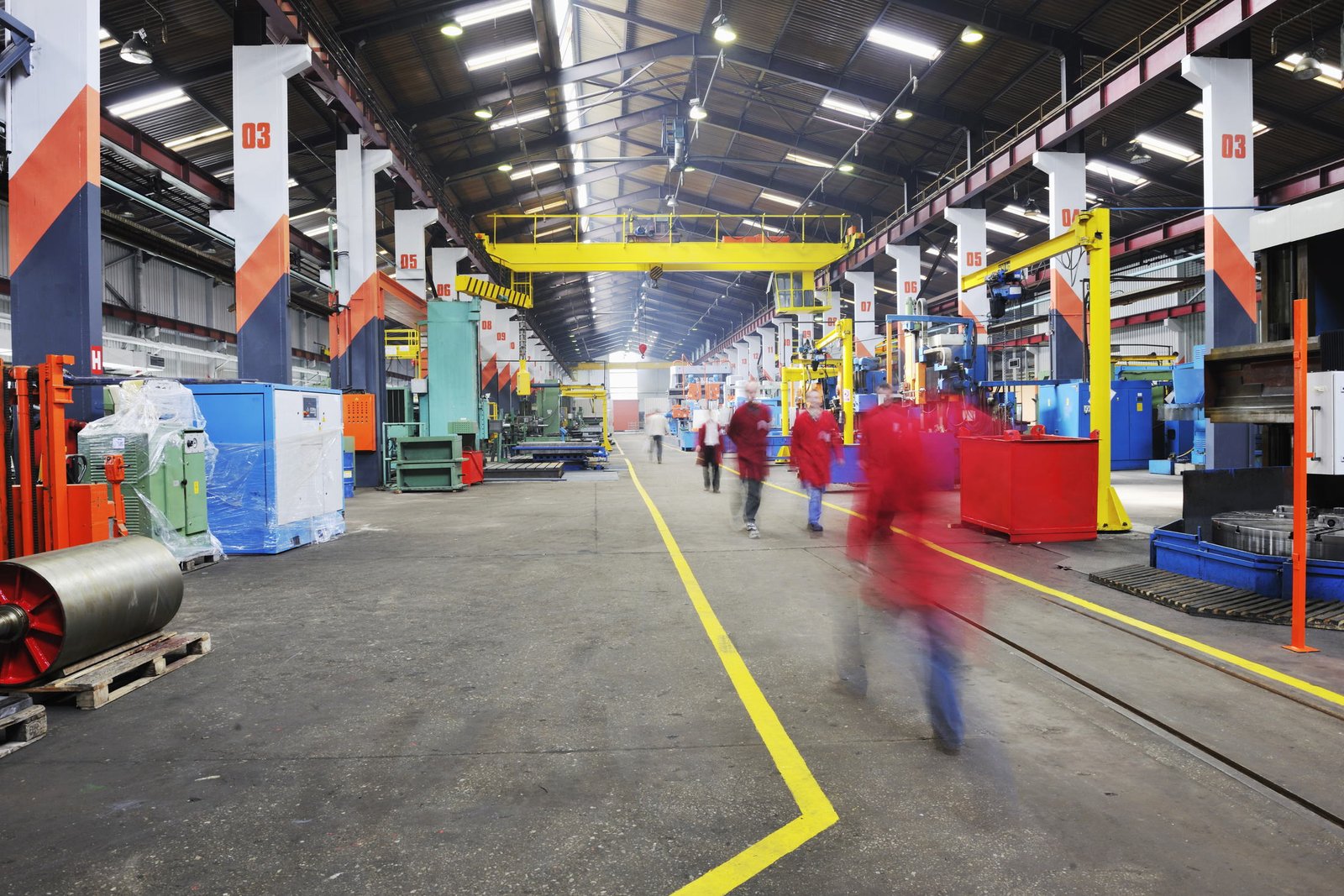 The main aisle of a working machine shop, floor markings underfoot and an overhead crane above.